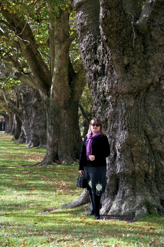 Mardi in foreground in black dress and pink scarf stanging in front of tree. Behinf her off at an angle are several other trees in line