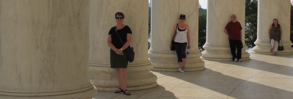 Image is of four people leaning against large ornate pillars. They are shaded and the sun streams through the gaps of the pillars.