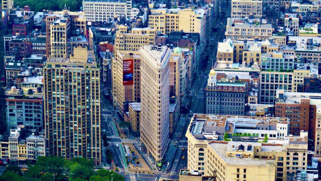Image of the Flatiron Building in NYC from above and a distance away. The flatiron thin edge faces forward and two streets fan out. Other buildings fill the frame.behind it.