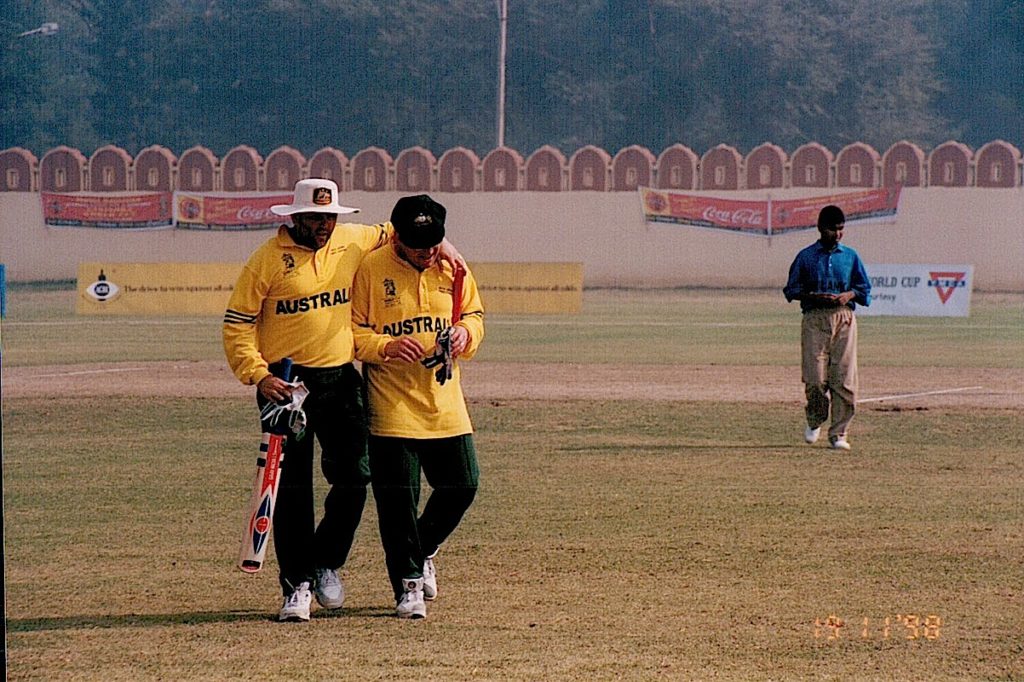 A picture of a cricket field, with me and Nejat in the foreground leaving the field. A lone Sri Lankan cricketer is in the background. The field is heavy with smog.
