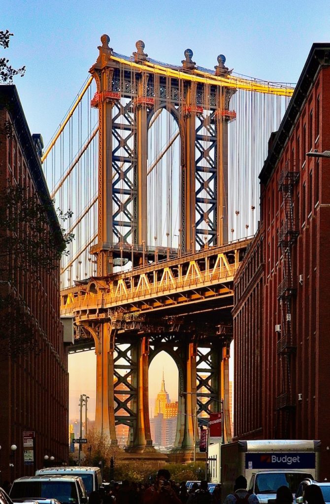 Image of Manhattan bridge between two buildings. In the distance the Empire Sate building stands under the arch of the bridge.