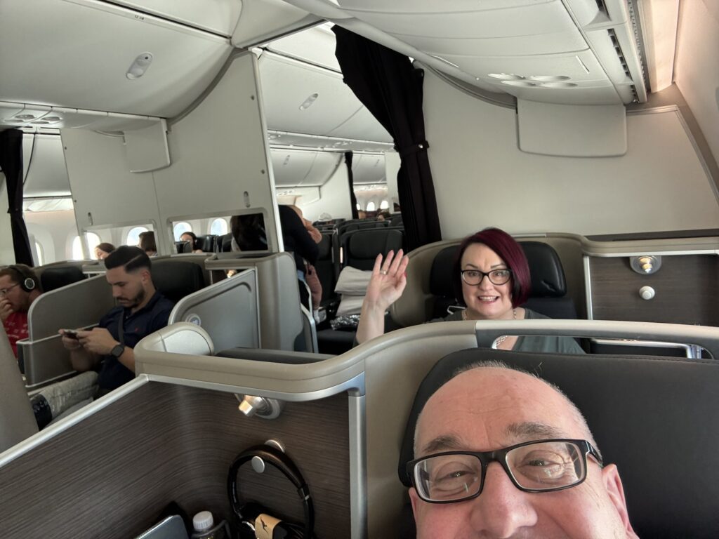 Michael is taking a selfie with himself and Mardi in the background in their seats on the 787 to New York