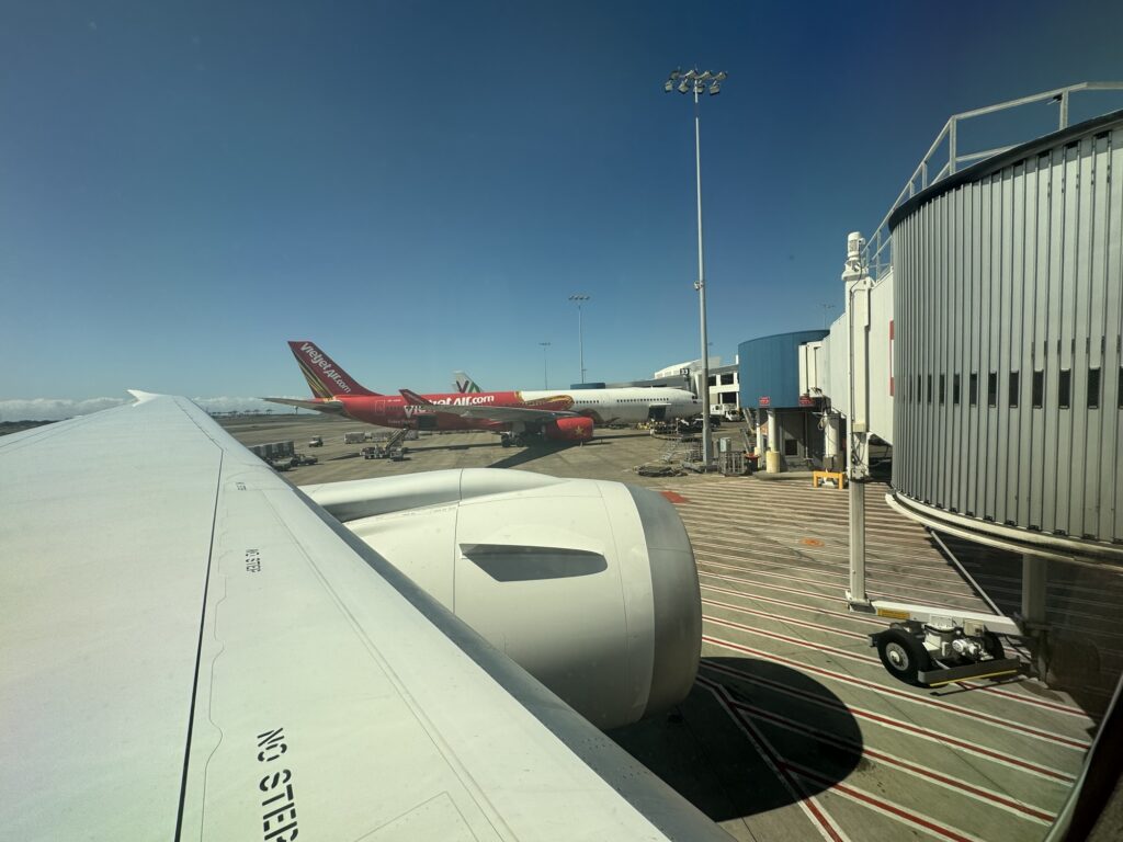 Wing and engine on the 787 as a taxi away from Sydney airport another aircraft is in thedistance. The sky is blue and it is a very sunny day