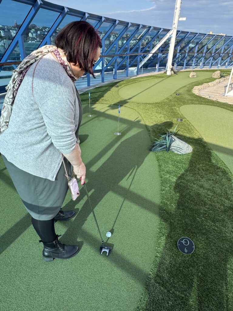 Mary putting on a green on the deck of insignia it’s bright and sunny. The hole is about 3 m from her. She’s about to take a stroke to the right as hazard.