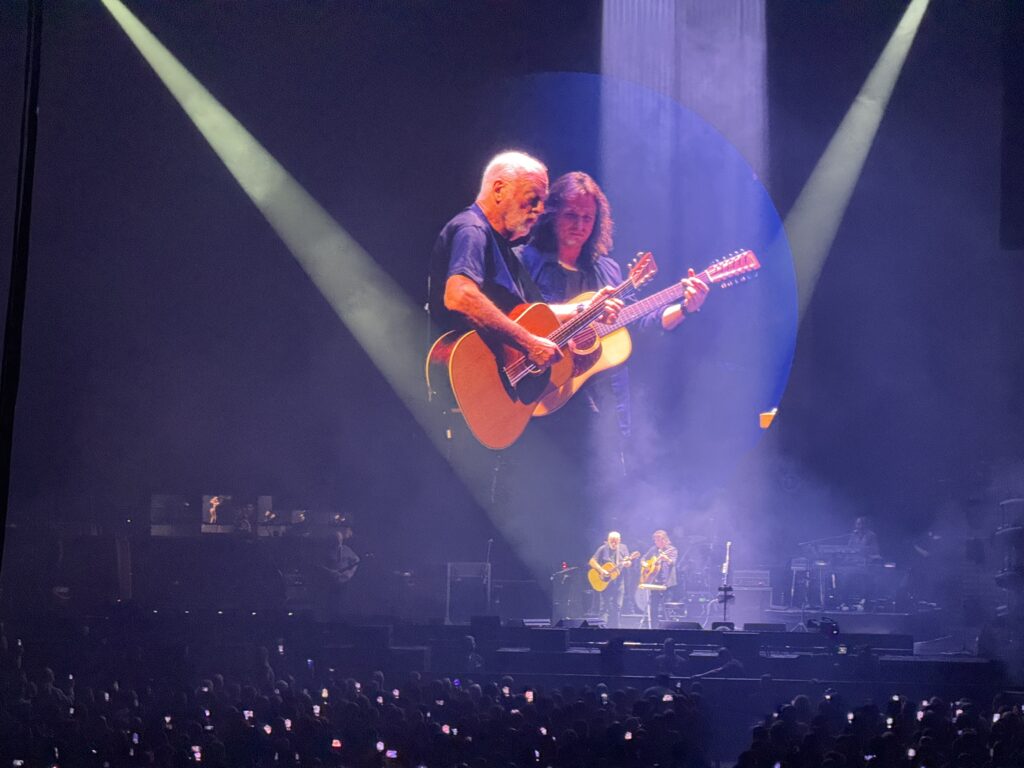 Gilmour projected on the massive screen. Black t shirt playing an acoustic guitar