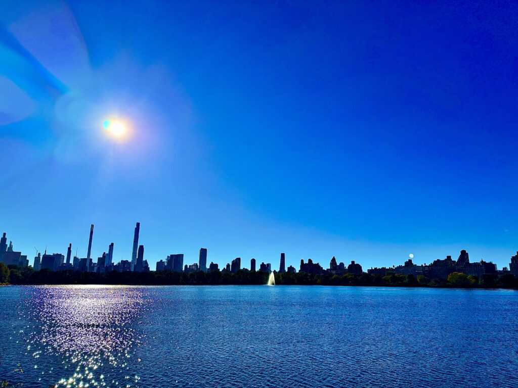 The sun, high in the blue sky over the reservoir. The skyline of manhattan dilohetted.