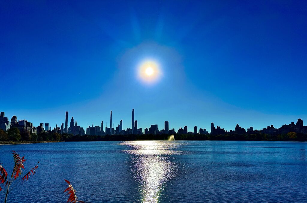 The sun, high in the blue sky over the reservoir. The skyline of manhattan dilohetted.