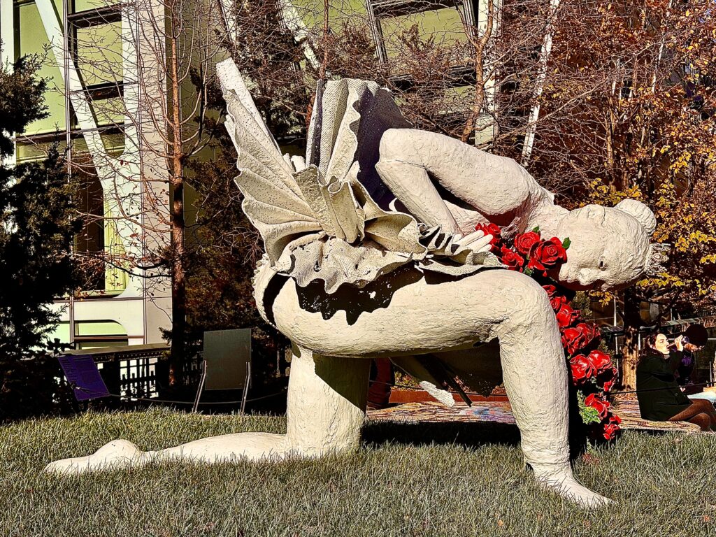 A white statue of a ballerina. She’s leaning forward on one knee. She’s holding a red rose.